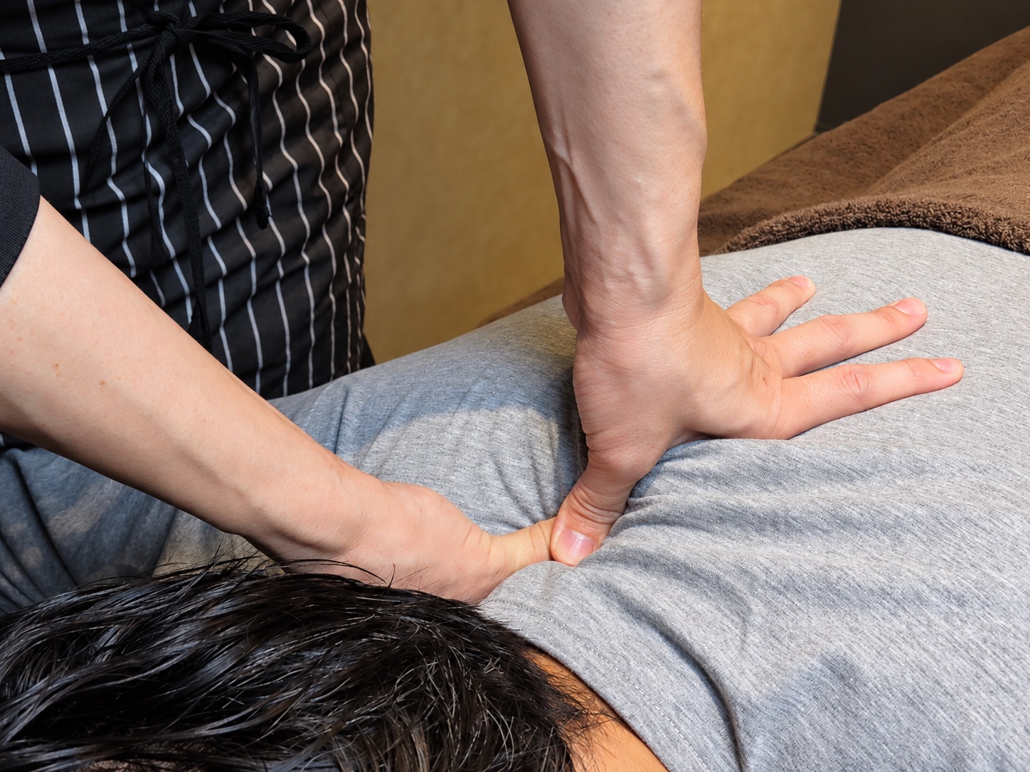 Hands applying sustained pressure with both thumbs to the neck and shoulders, over a grey top